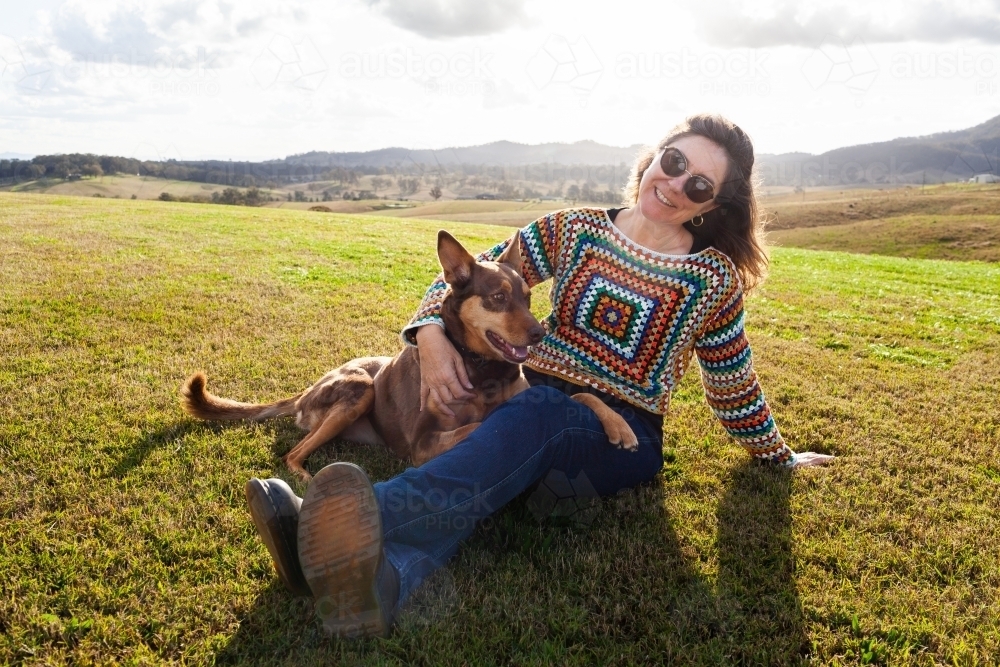 Companions - happy middle age woman and kelpie dog sitting together on farm hilltop - Australian Stock Image