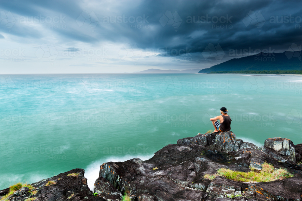 Contemplative young man sitting on a rock, looking at stormy ocean seascape and tropical coastline - Australian Stock Image