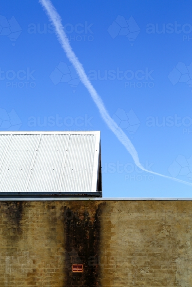 Contrails, a brick wall, and tin roof under a vivid blue sky. - Australian Stock Image