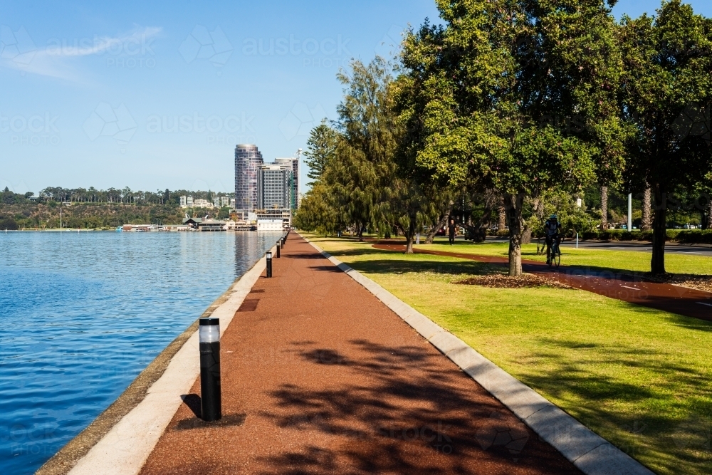 Converging lines of orange path, path lights and trees beside blue water with building and blue sky - Australian Stock Image