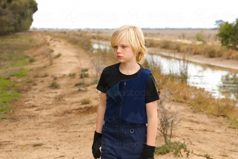 Image of Country boy wearing overalls standing with serious expression by  channel on farm - Austockphoto