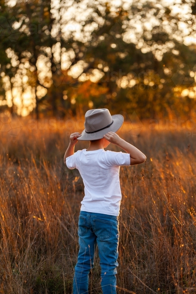 country kid putting hat on in paddock looking away - Australian Stock Image