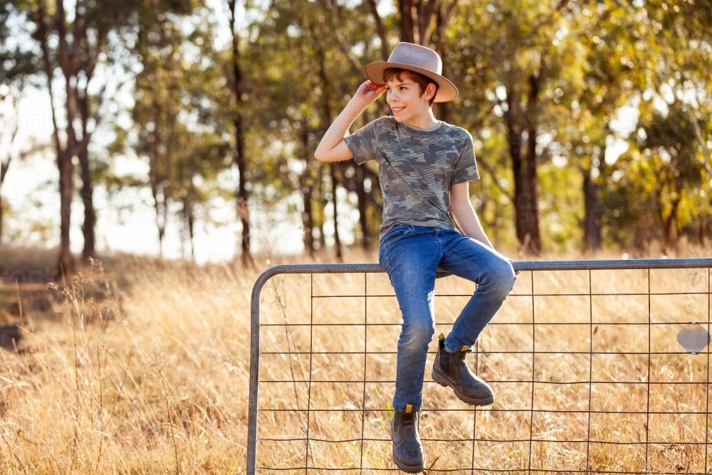 Country preteen kid sitting on paddock fence with hand up to hat - Australian Stock Image