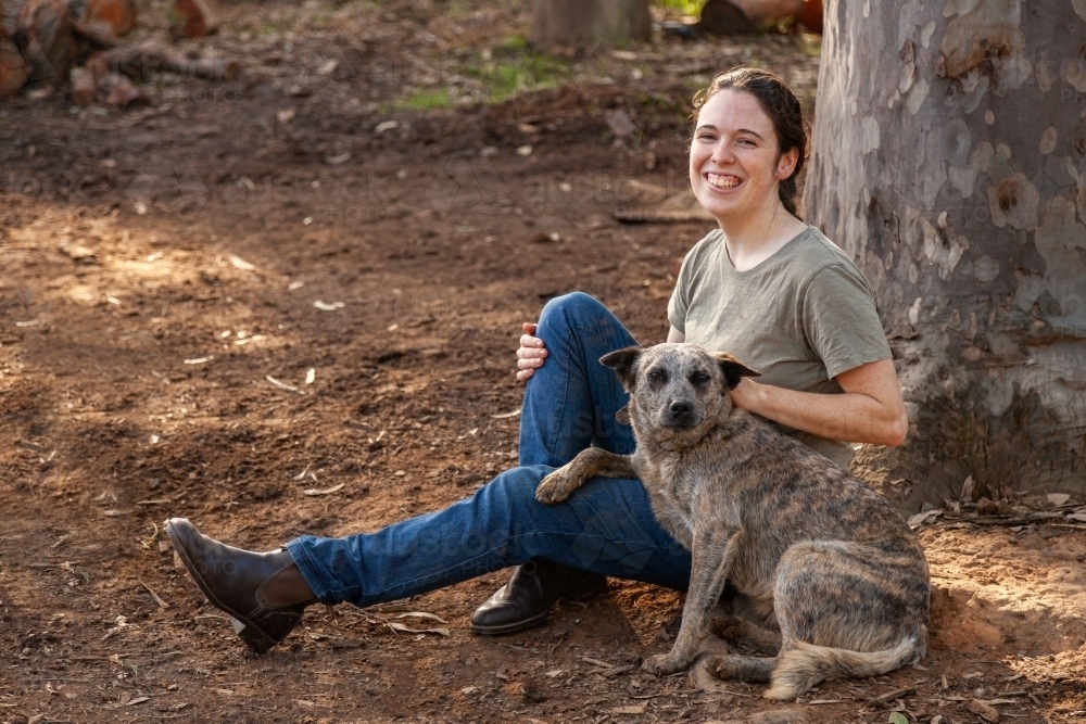 Country woman sitting beside gum tree with pet dog - Australian Stock Image