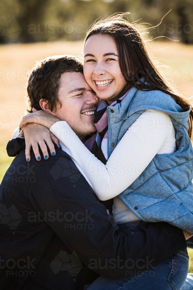 Couple cuddling with girlfriend sitting on boyfriend's lap with arms around neck laughing - Australian Stock Image