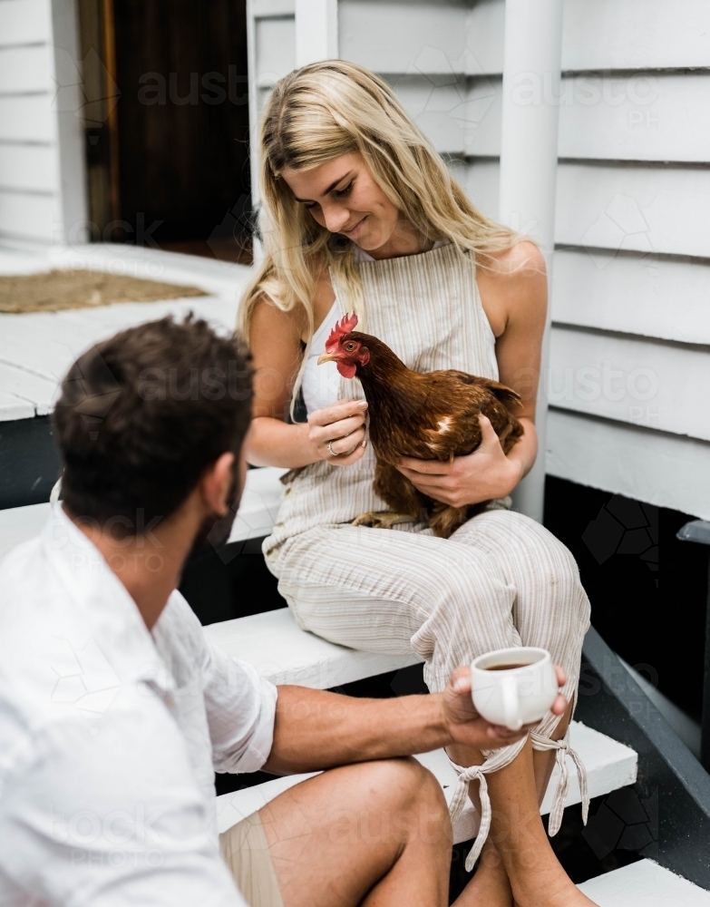 Couple having a cup of coffee on steps of verandah, with woman holding a chicken. - Australian Stock Image