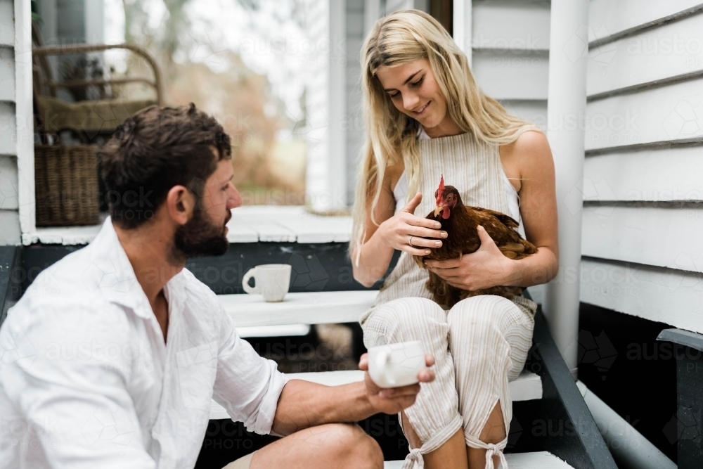 Couple having a cup of coffee on steps of verandah, with woman holding a chicken. - Australian Stock Image