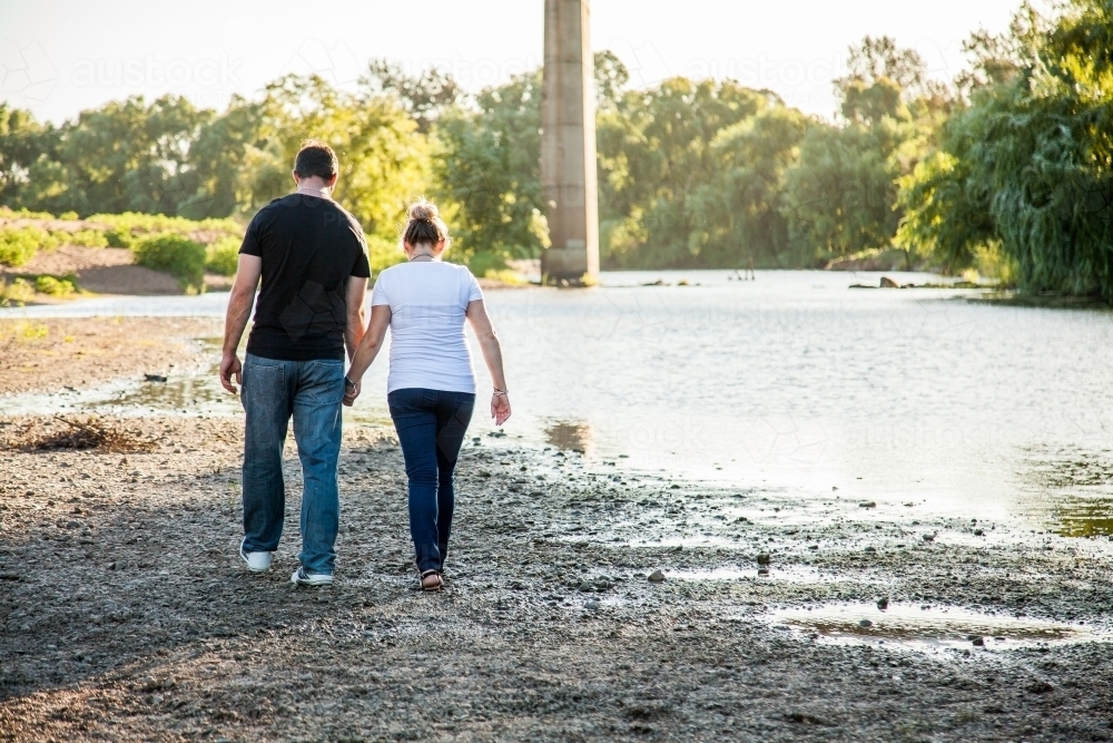 Couple holding hands walking away along river - Australian Stock Image