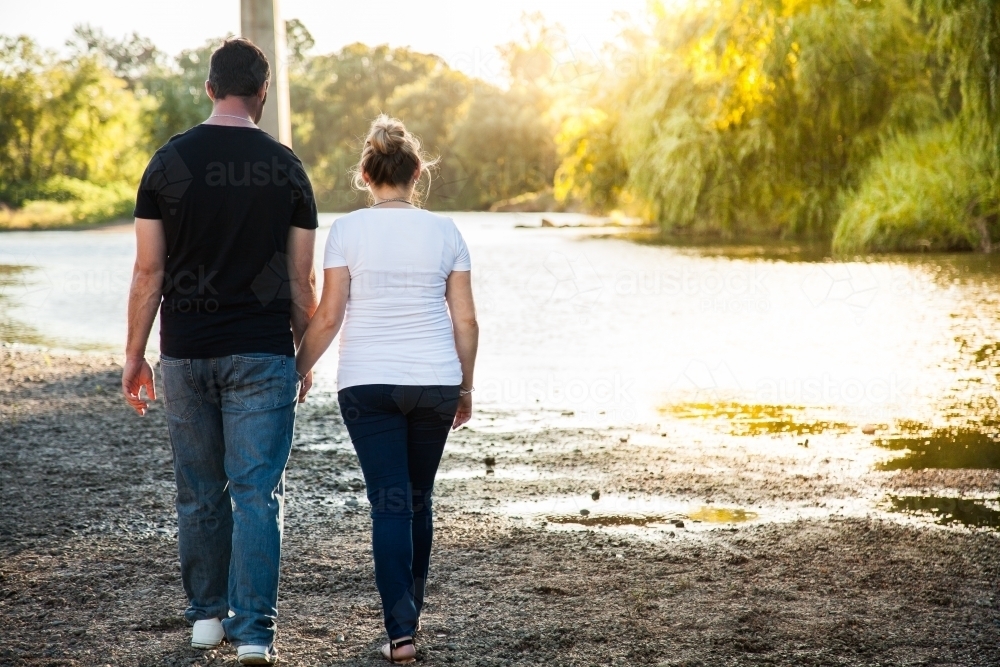 Couple holding hands walking away along river - Australian Stock Image