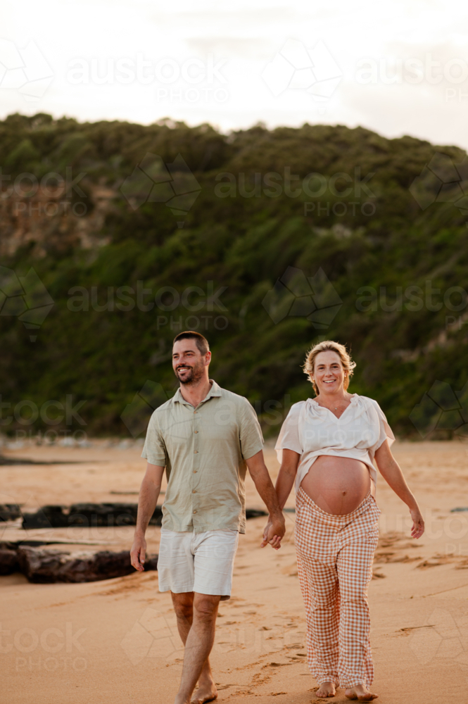 Couple holding hands walking on beach at sunset, pregnant woman in relaxed clothing - Australian Stock Image