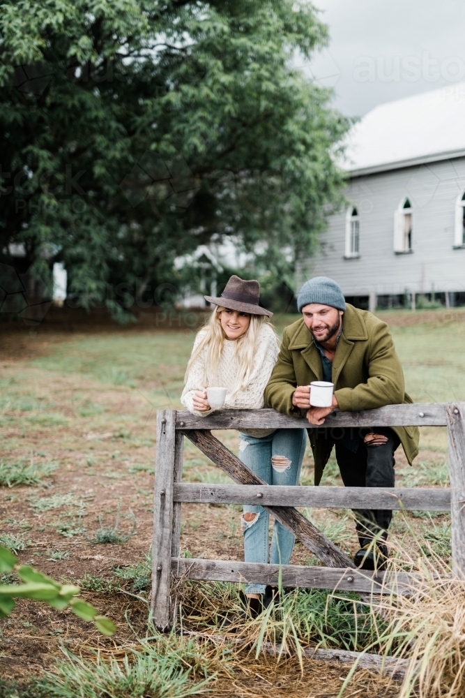 Couple leaning on farm fence with farmhouse in background - Australian Stock Image