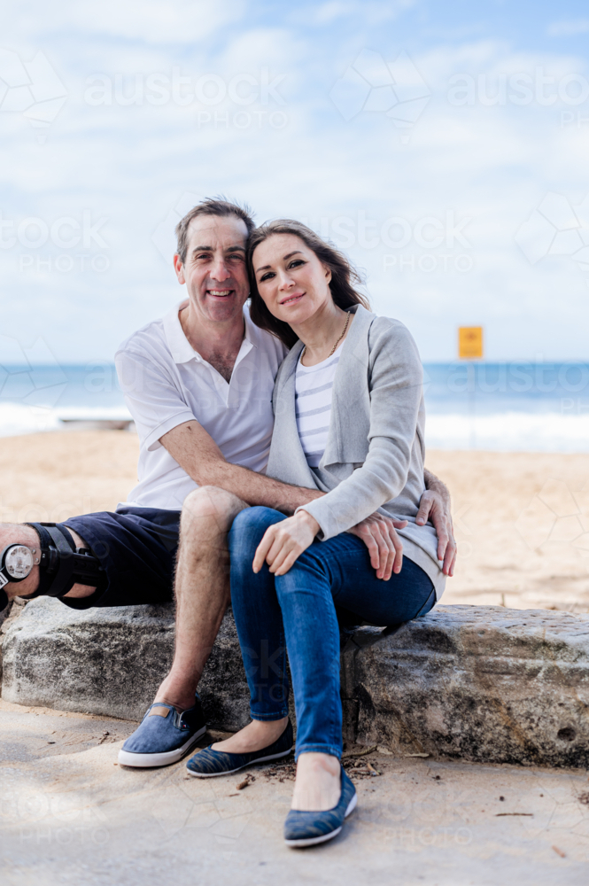 Couple relaxes on a rocky shoreline, smiling warmly against a scenic beach backdrop - Australian Stock Image