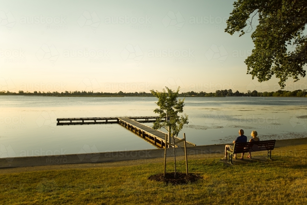 Couple sitting on bench watching the sunset over a lake - Australian Stock Image