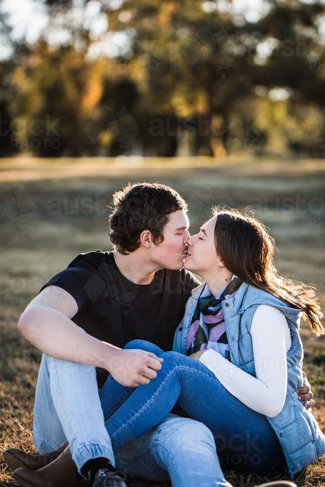 Couple sitting on grass with legs entwined kissing - Australian Stock Image