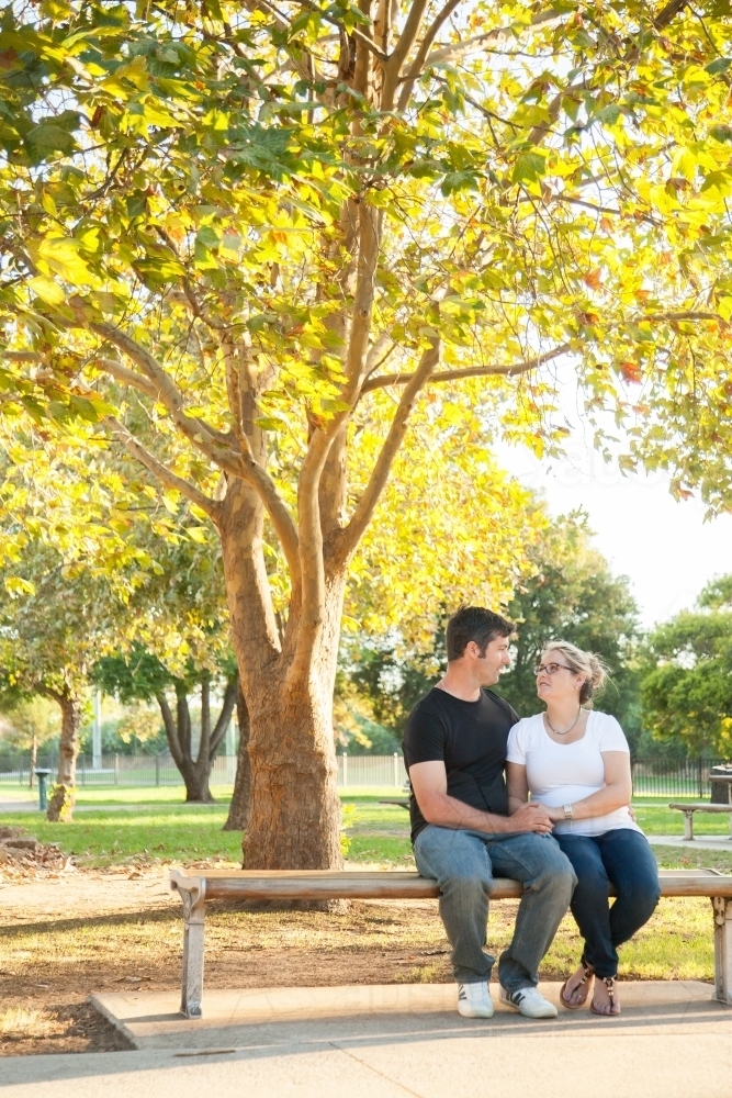 Couple sitting on park bench under autumn tree looking at each other - Australian Stock Image