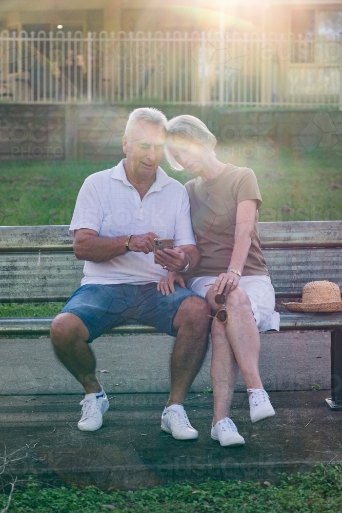 couple sitting on park bench, using phone - Australian Stock Image