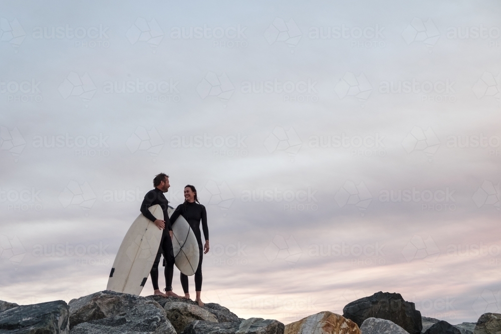 Couple standing on coastal rocks wearing wetsuits and carrying surfboards looking at sunset - Australian Stock Image