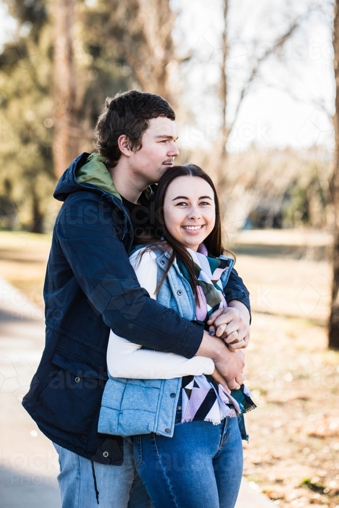 Couple standing with man's arms wrapped around woman holding hands looking off into distance - Australian Stock Image