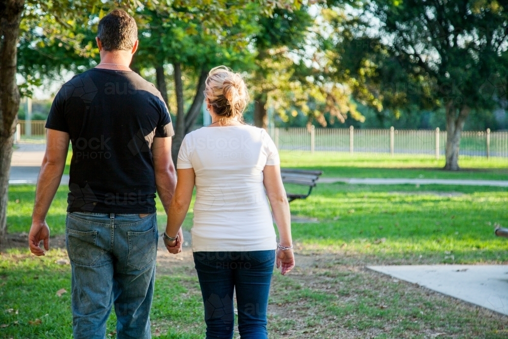 Couple walking away through park holding hands - Australian Stock Image