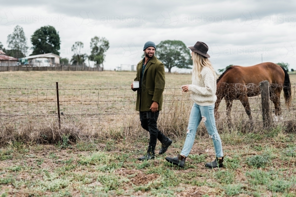 Couple walking past horses on a farm, holding cups of coffee : Austockphoto Couple walking past horses on a farm, holding cups of coffee - Australian Stock Image