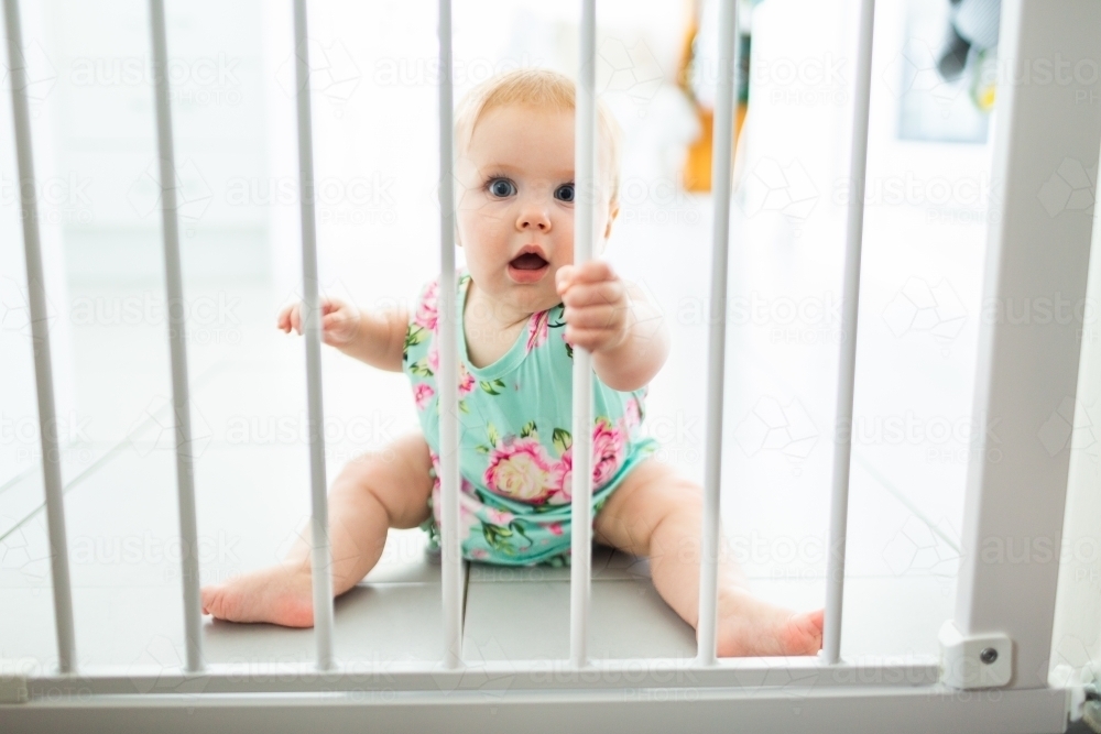 Crawling baby in kitchen holding onto bars of safety baby gate - Australian Stock Image