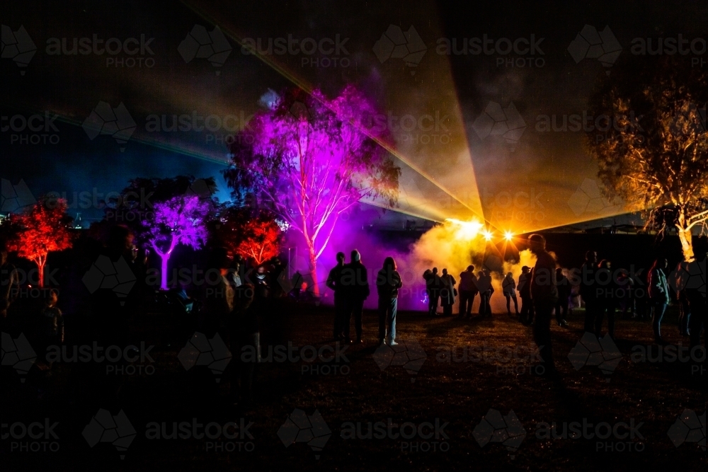 Crowd of people watch night time laser light display beams of colourful light shining on fog - Australian Stock Image