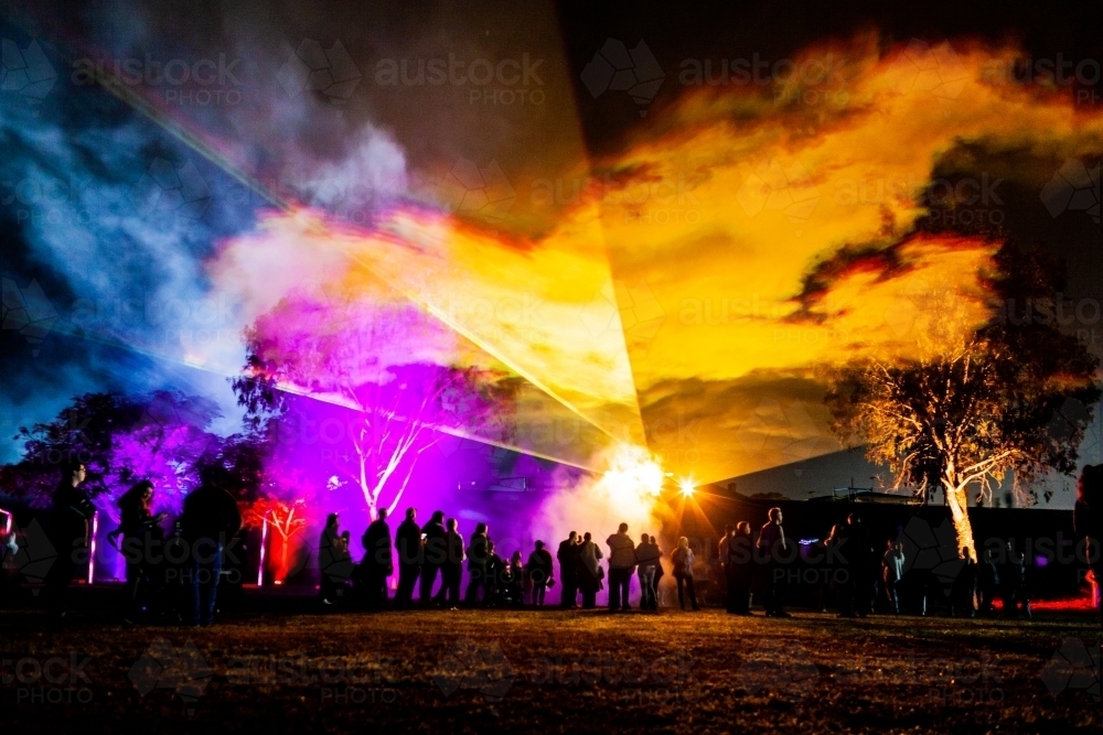 Crowd of people watch night time laser light display beams of colourful light shining on fog - Australian Stock Image