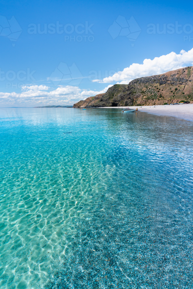Crystal clear turquoise sea water along pebble beach with coastal cliffs under blue sky - Australian Stock Image