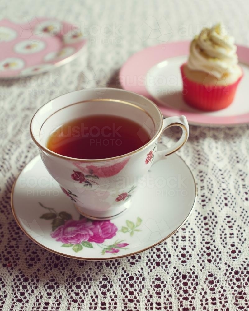 Cup of tea in vintage floral cup and saucer with cupcake behind - Australian Stock Image