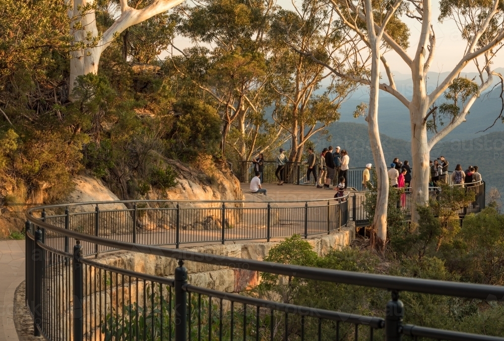 Curved concrete pathway through bushland, with safety fence, gumtrees and distant walkers and view - Australian Stock Image