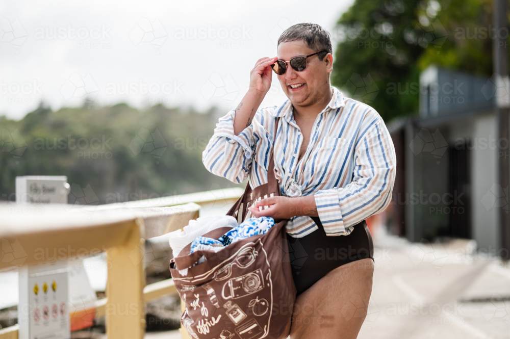 Curvy woman standing on coastal path wearing sunglasses carrying a brown beach bag on sunny day - Australian Stock Image