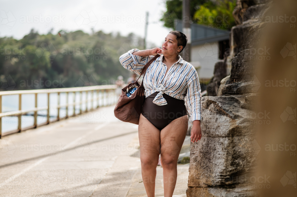 Curvy woman walking on coastal path carrying a brown beach bag on sunny day - Australian Stock Image
