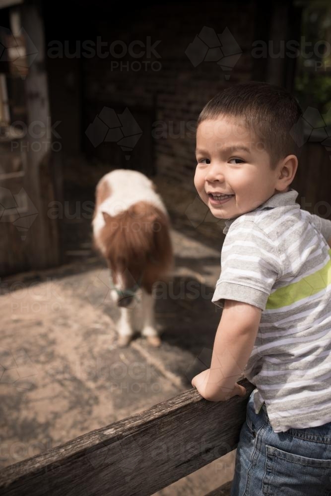 Cute 2 year old mixed race boy plays on a fence in an animal stable - Australian Stock Image