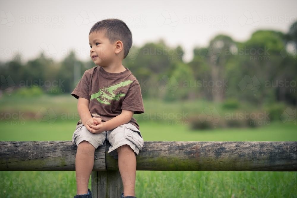 Cute 3 year old boy sits on a wooden log fence in a suburban park - Australian Stock Image