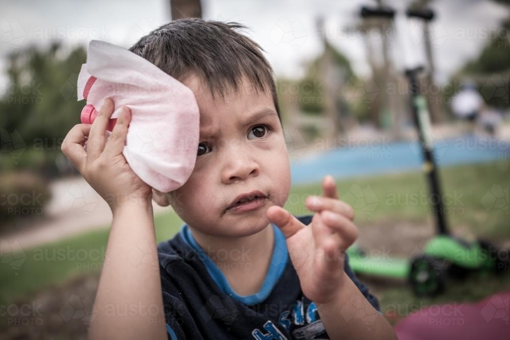 Cute 3 year old mixed race boy holds an esky ice pack on his head after falling off his scooter - Australian Stock Image