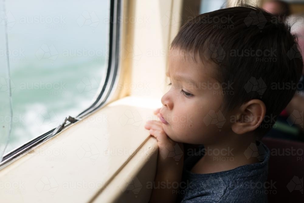 Cute 3 year old mixed race boy looks out a boat window - Australian Stock Image