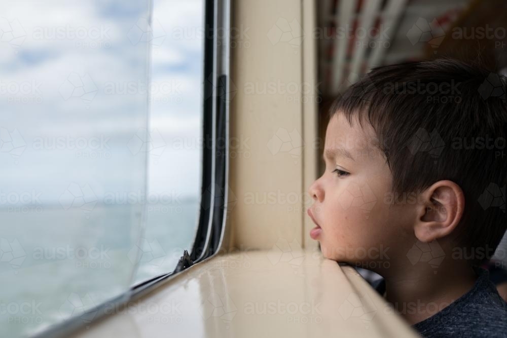 Cute 3 year old mixed race boy looks out a boat window - Australian Stock Image