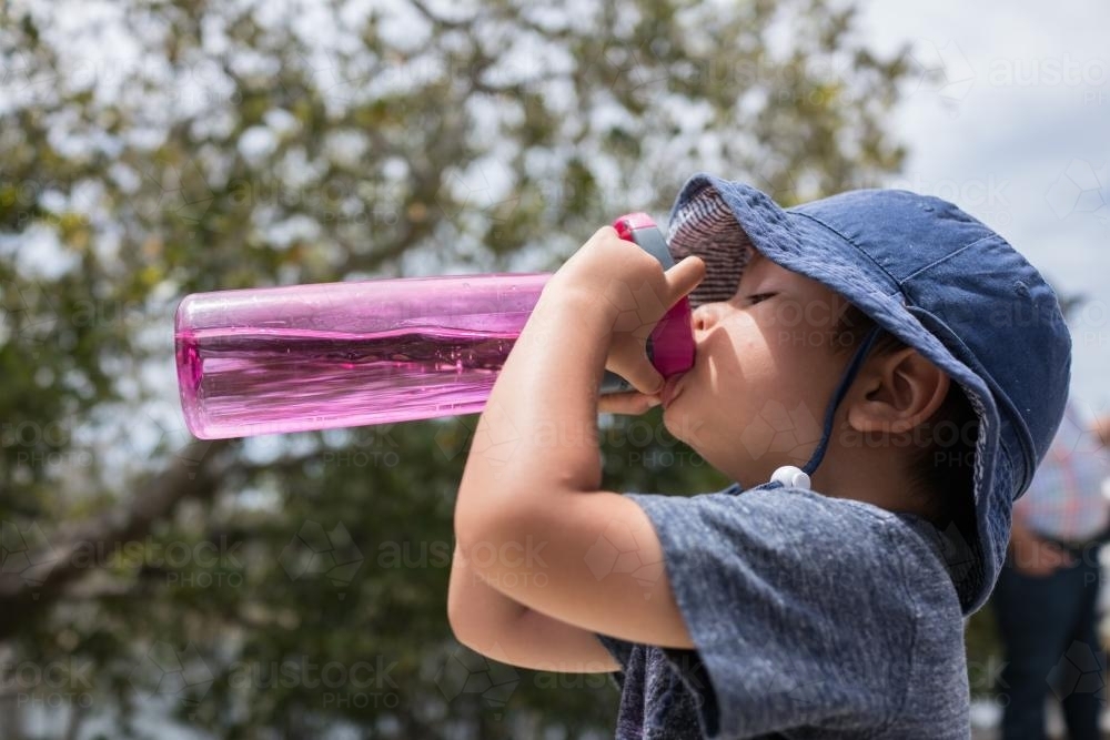 Cute mixed race boy drinks from a pink water bottle outside in the sun - Australian Stock Image