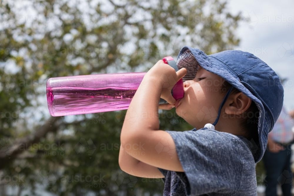 Cute mixed race boy drinks from a pink water bottle outside in the sun - Australian Stock Image