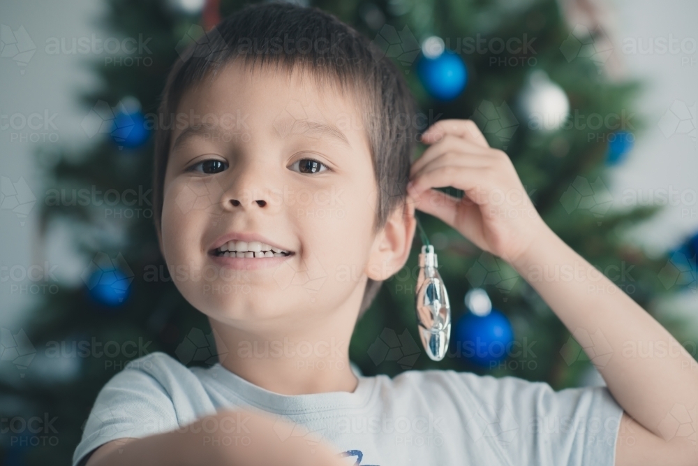 Cute mixed race boy hanging Christmas tree decorations on ear - Australian Stock Image