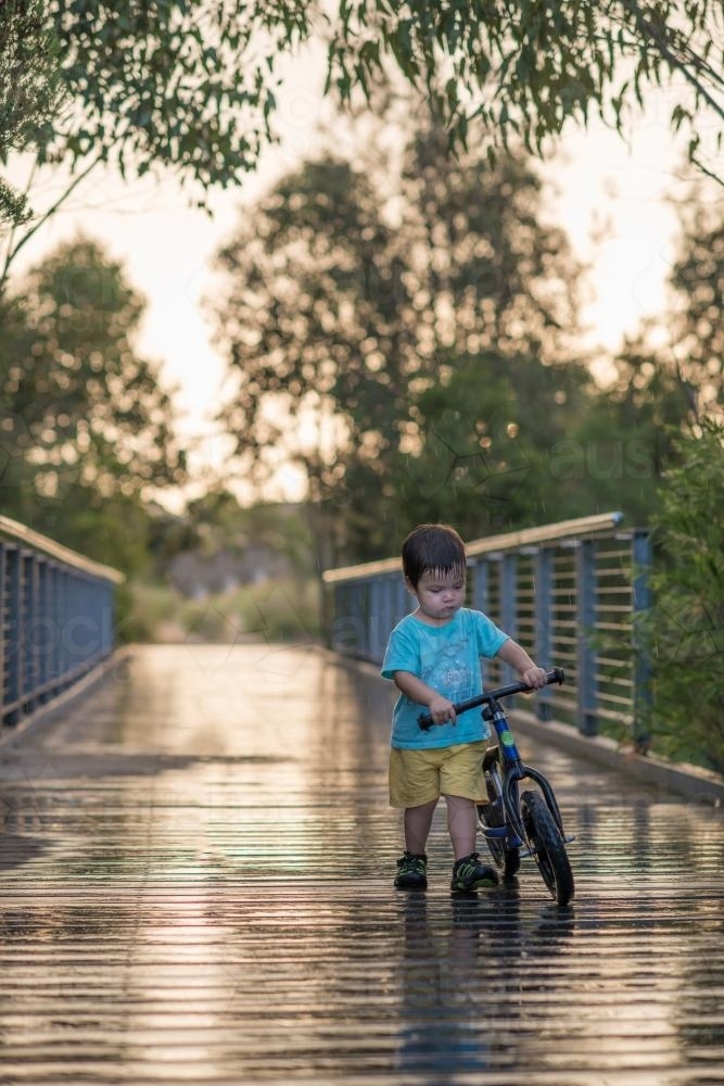 Cute mixed race boy walks home with his bike in the rain - Australian Stock Image