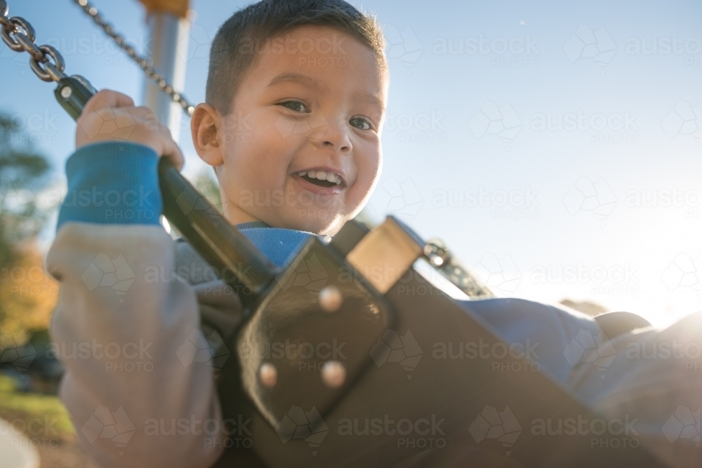 Cute mixed race three year old boy playing on a swing in a suburban playground - Australian Stock Image