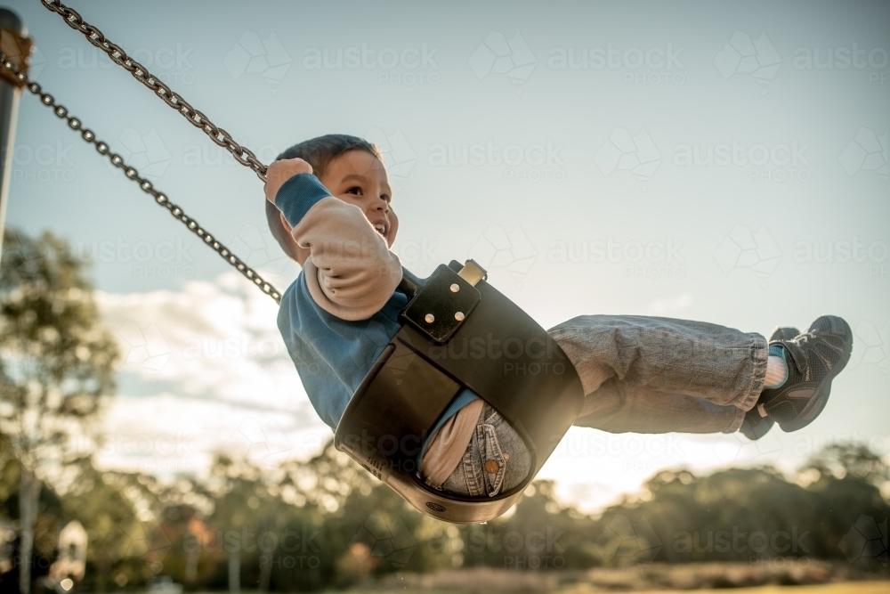 Cute mixed race three year old boy playing on a swing in a suburban playground - Australian Stock Image