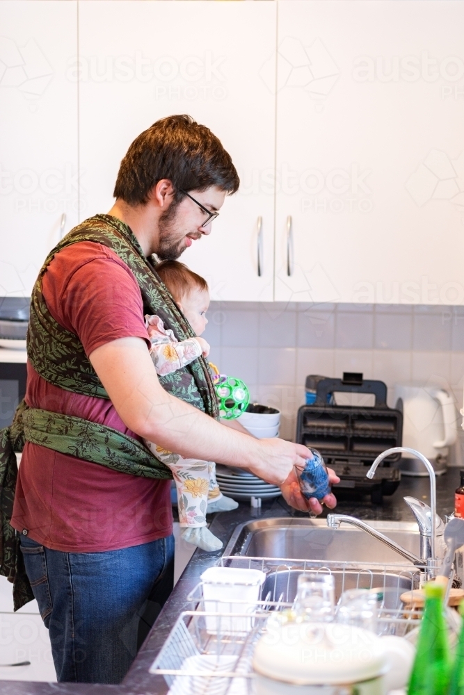 Dad in kitchen doing dishes while holding baby in wrap - Australian Stock Image