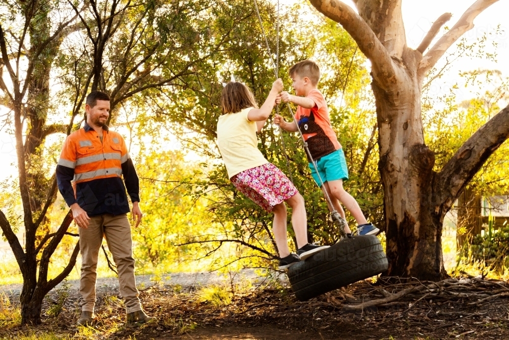 Dad playing with children in backyard on tyre swing in the afternoon - Australian Stock Image