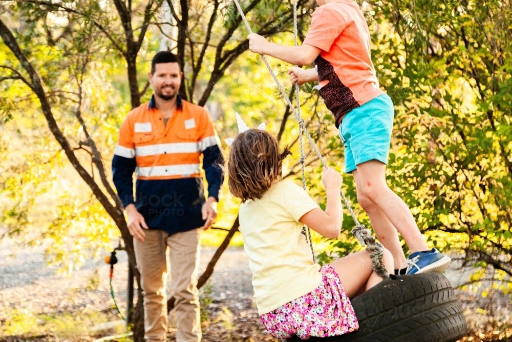Dad playing with children in backyard on tyre swing in the afternoon - Australian Stock Image