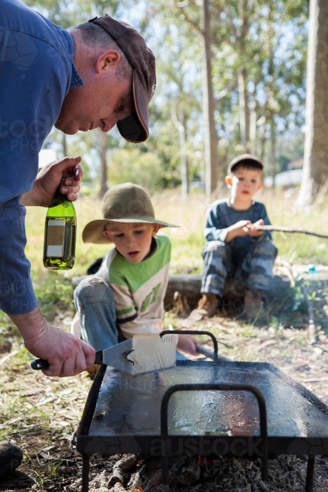 Dad preparing hot plate for cooking over campfire while boys watch - Australian Stock Image