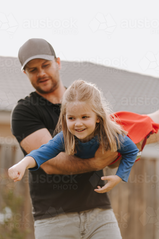Dad simulating a flight for daughter in superhero costume. - Australian Stock Image