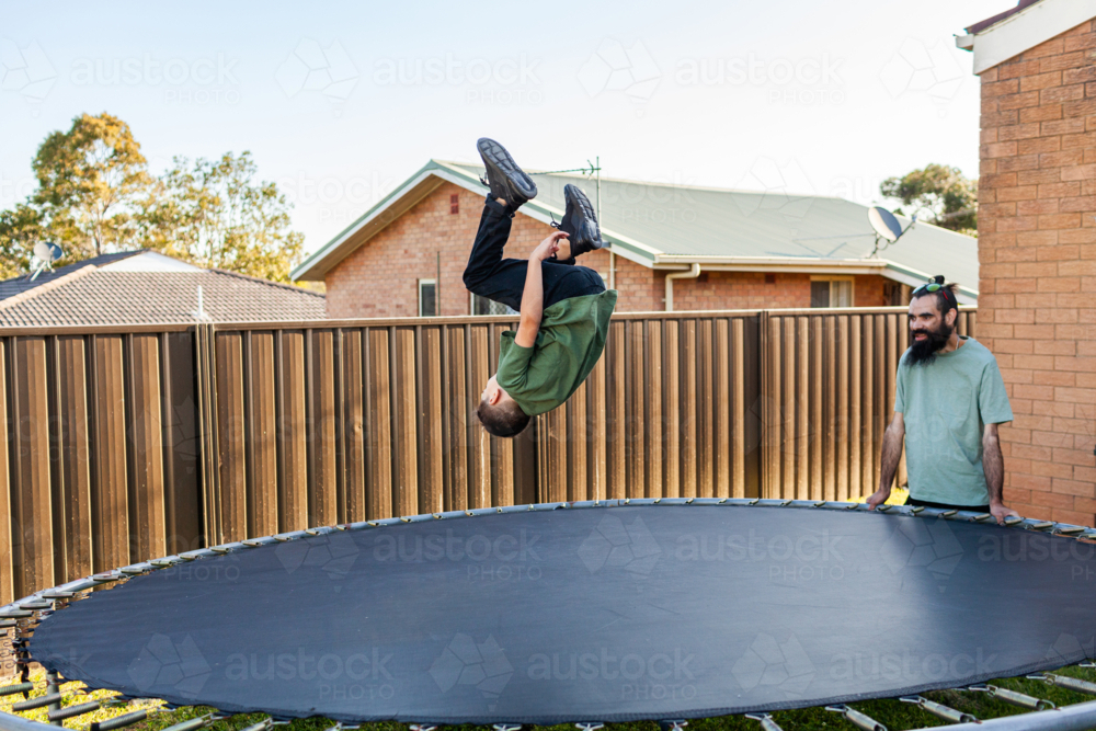 Dad watching eight year old Aussie kid jumping and doing flips on trampoline in backyard - Australian Stock Image