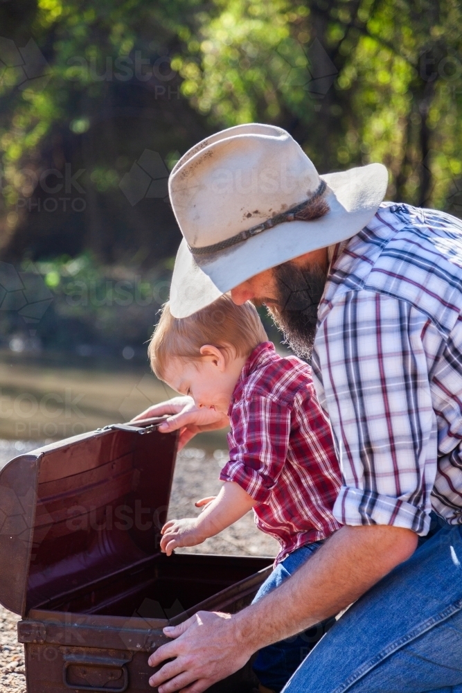 Dad with son opening treasure chest beside river - Australian Stock Image